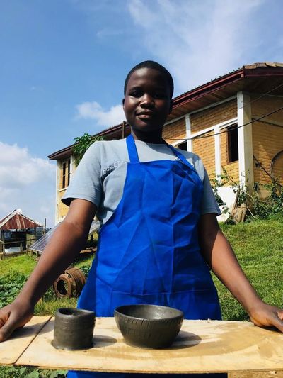 A student in a blue apron presenting two pots she has made, with school buildings and solar panels behind