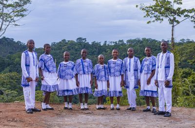 Eight Mfàh'Mfàh Academy students in blue-and-white uniform standing together on a ridge above rainforest